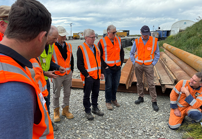 Group of people in high-visibility orange vests observing a worker kneeling on the ground at an outdoor work site with pipes and equipment in the background.