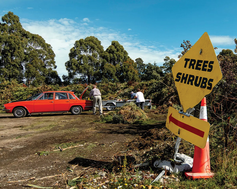 A red vintage car is parked near a trailer in an outdoor area with trees and bushes. Two people are working near the trailer. In the foreground, there's a yellow sign with "TREES SHRUBS" written on it, and an arrow pointing right.