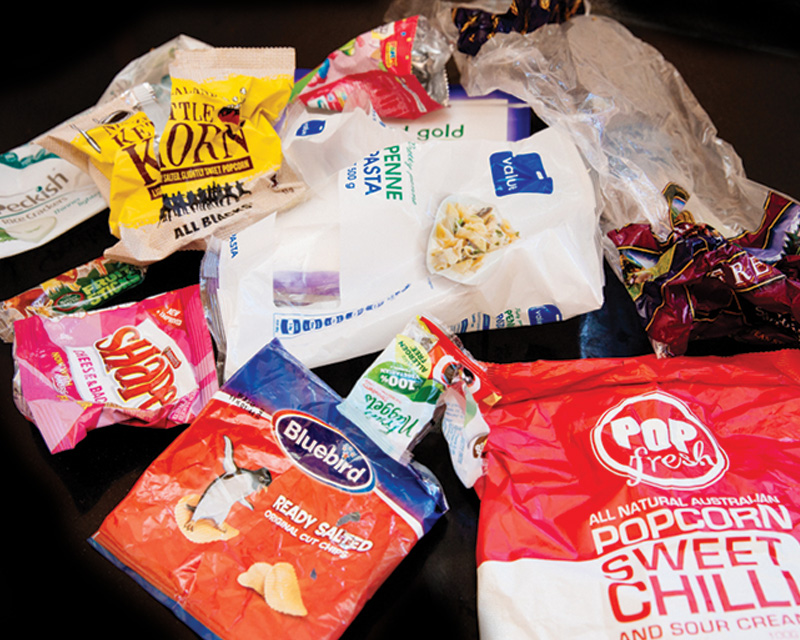 A scattered collection of various colorful, empty snack and food packaging, including popcorn, chips, candy, and pasta bags