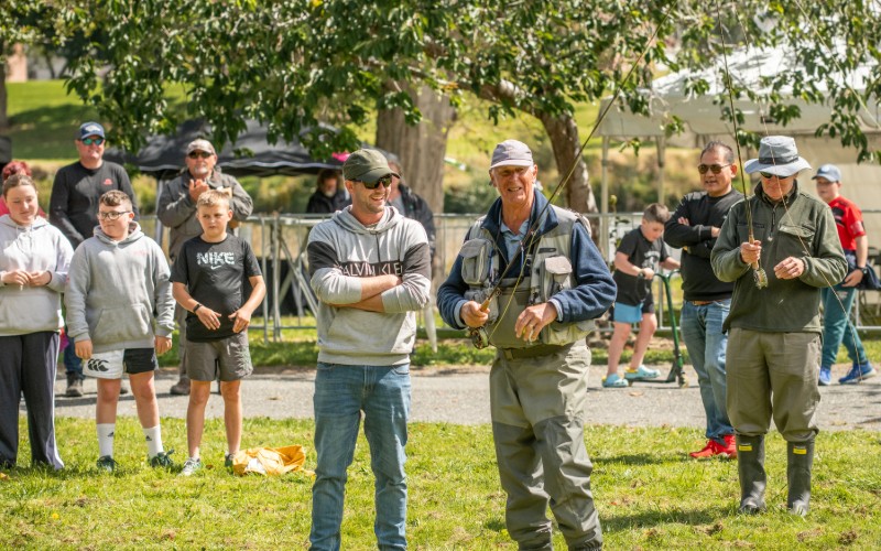 Fishing guide teaching a man to cast at On the Fly Festival by the Mataura River. There is a crowd of people watching. 